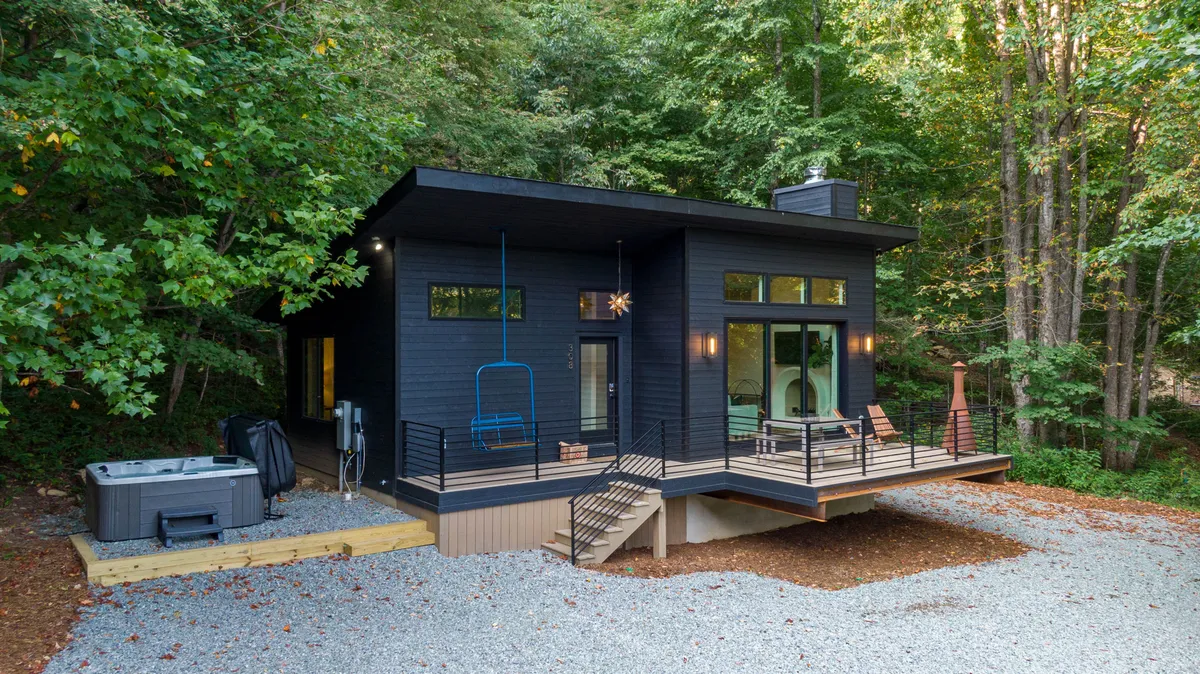 The hero image features a sleek, modern black cabin nestled among tall trees with a private deck. The inviting hot tub and comfortable outdoor seating area are focal points, surrounded by nature, creating an atmosphere of tranquility and seclusion. The deck offers scenic views, ideal for sunsets and stargazing.