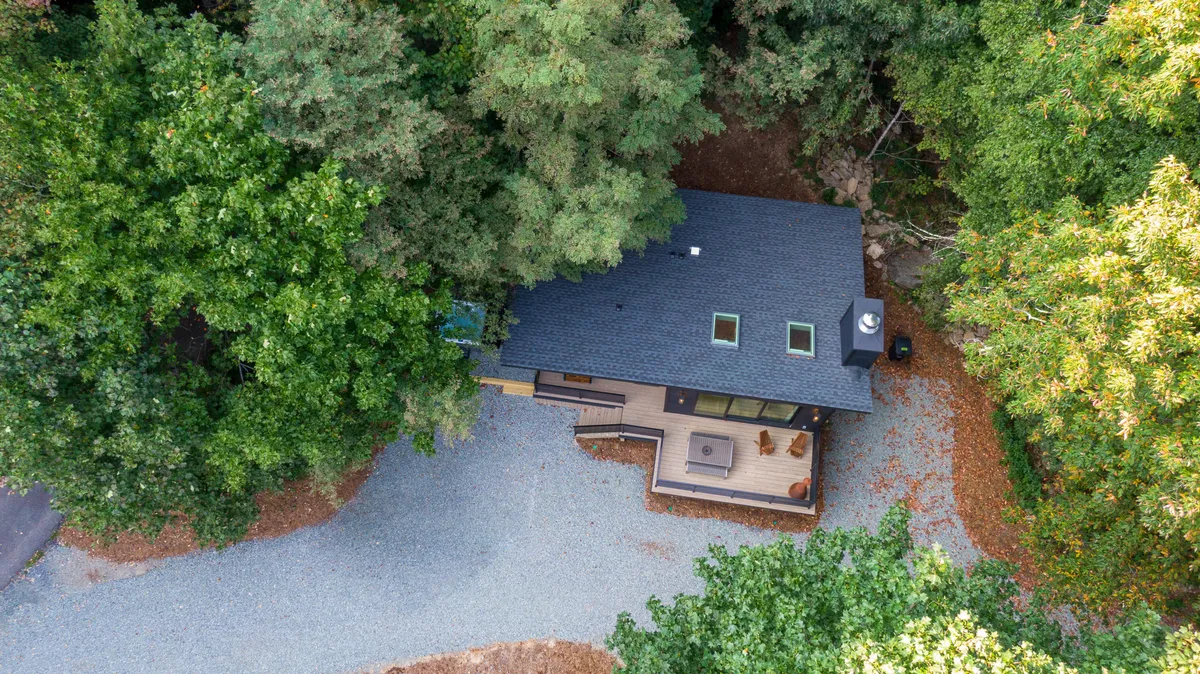 This photo shows the house from another angle, featuring an additional outdoor seating area, a gravel driveway, and mature trees that envelop the property. The peaceful setting is ideal for morning coffee or evening stargazing, and the open, accessible exterior conveys a sense of space for both guests and their dogs to enjoy.
