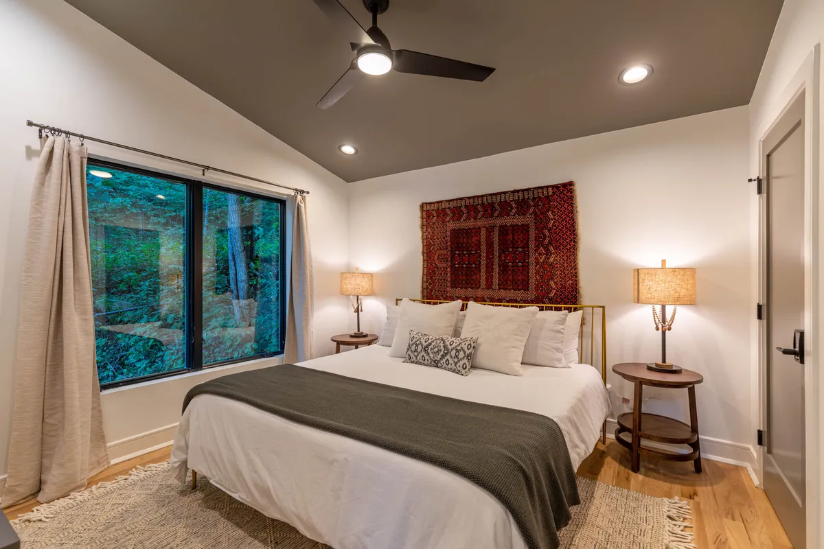 This image features a serene bedroom setting with a large bed adorned with decorative pillows, flanked by wooden side tables with lamps. Ample natural light streams in from expansive windows that offer tranquil nature views. The room is accented by a textured wall hanging that adds a touch of warmth and personality.