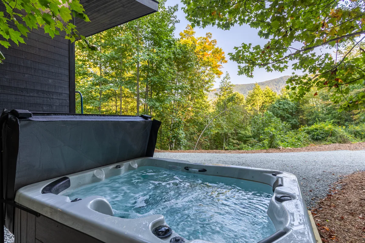 A serene outdoor scene featuring a four-person hot tub surrounded by tall trees, with a sweeping mountain view in the background. The deck is spacious, providing a sense of seclusion and luxury with nature just steps away. The lighting suggests the possibility of sunset or early evening soaks, with the hot tub as the focal point.