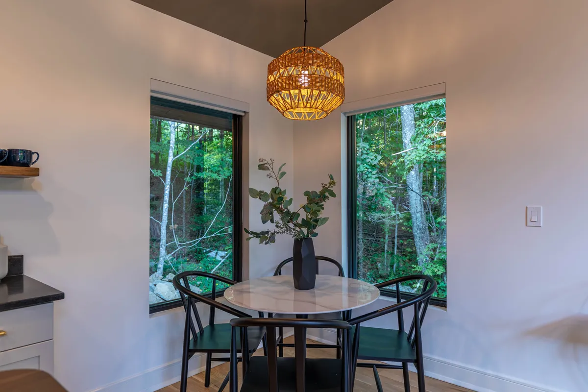 An intimate round dining table set beside large windows, adorned with a woven pendant light and simple greenery in a vase. The windows open to lush outdoor views, setting a tranquil, cozy mood perfect for quiet mornings or romantic dinners.