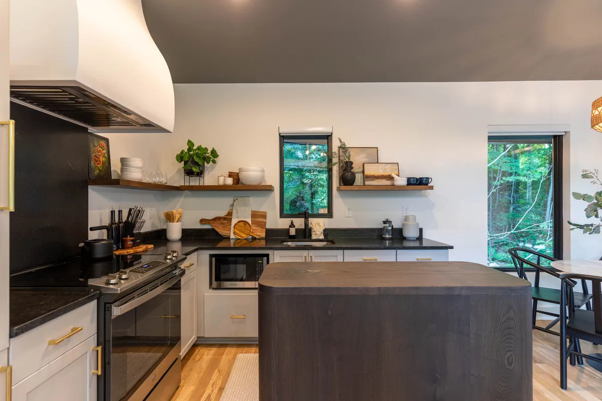 A bright, welcoming kitchen scene centered on a large island with black countertops, wooden shelves stocked with kitchenware, and lush plants adding warmth. Large windows offer a hint of the surrounding views and natural light floods the space, highlighting the kitchen as a gathering spot.
