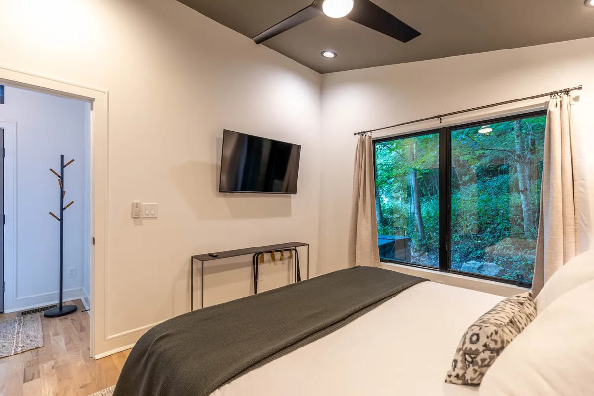 This image shows a comfortable bed with a sleek black throw blanket, a large window showcasing outdoor views, and a smart TV mounted on the wall. The neutral palette and calm ambiance suggest a relaxing and modern bedroom environment.