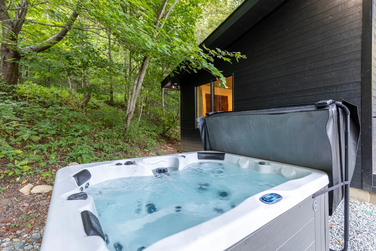 The exterior of the cabin features a private hot tub set among tall trees, with the back of the house visible. The ambiance is serene, inviting relaxation and intimate moments while being immersed in nature.