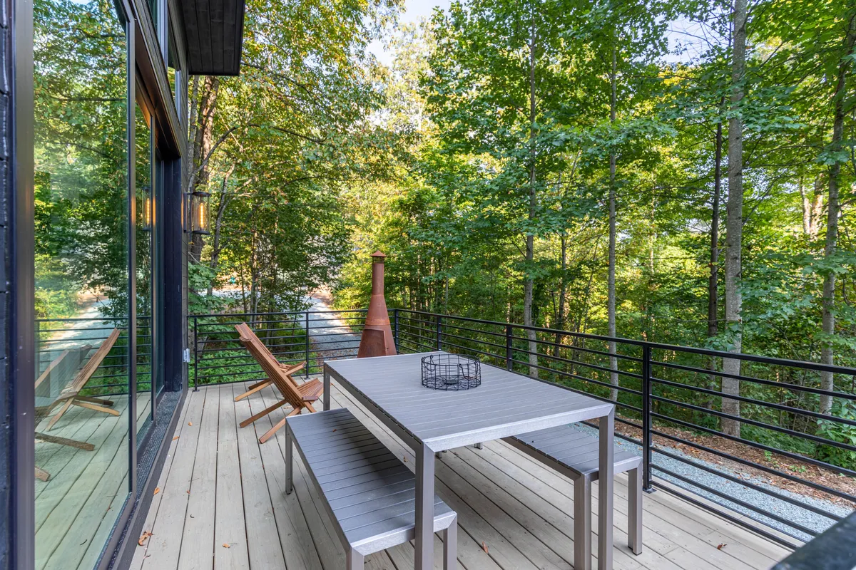 An inviting outdoor dining area set on a wooden deck, surrounded by mature trees and gentle sunlight. The dining table is large enough for group feasts, and a fire pit adds warmth and ambiance for evening gatherings. The space is both functional and picturesque, emphasizing alfresco dining and relaxation.