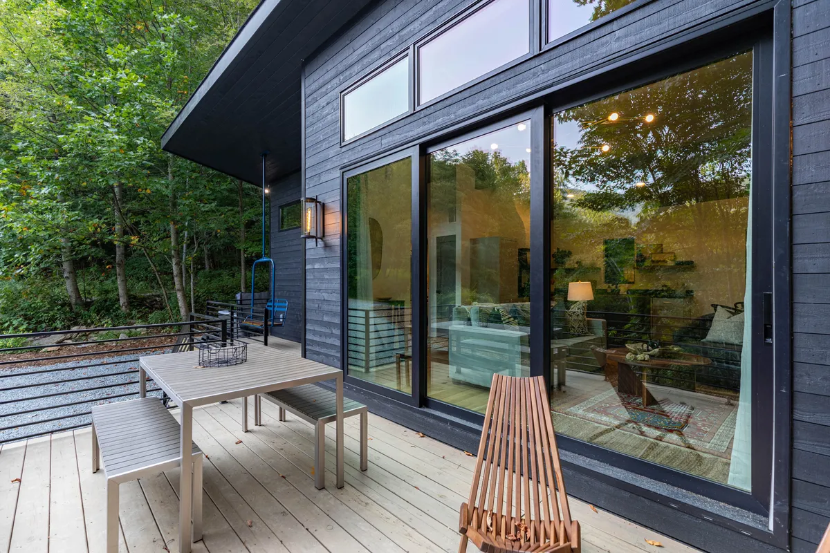 A tranquil deck scene with an outdoor dining table and wooden bench, surrounded by greenery and large glass windows reflecting the environment. The space is perfect for al fresco meals or enjoying a peaceful moment with a loved one.