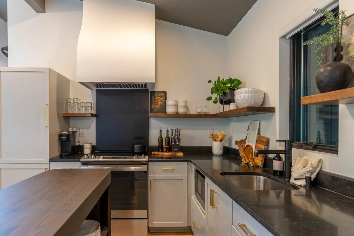 A contemporary kitchen featuring a central island with black countertop, stainless steel appliances including a refrigerator and oven, and sleek shelving with decorative touches. The layout invites both cooking and socializing, with clear visibility of the kitchen's central elements.