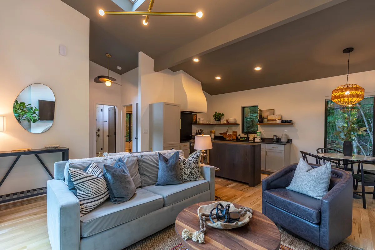 A sunlit living area featuring a plush sofa with decorative pillows, a wooden coffee table, and a stylish light fixture. The dining area is elegantly visible in the background, emphasizing the seamless flow between lounging and dining spaces. Ample windows bathe the space in natural light, enhancing feelings of warmth and welcome.