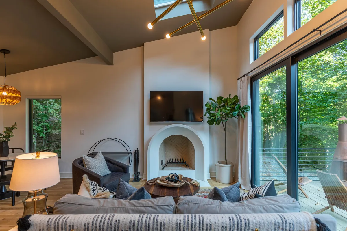 A bright, inviting living area with a modern fireplace as the centerpiece, large windows framing lush greenery, comfortable seating, and a stylish light fixture overhead. The space feels serene and connected to nature, with daylight streaming in and a tranquil atmosphere suited for both work and relaxation.